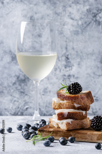 Grilled sandwich with melted goat cheese, blackberry, blueberry, rosemary and honey, served on wooden serving board with glass of cold white wine over gray blue texture background. Summer appetizer.