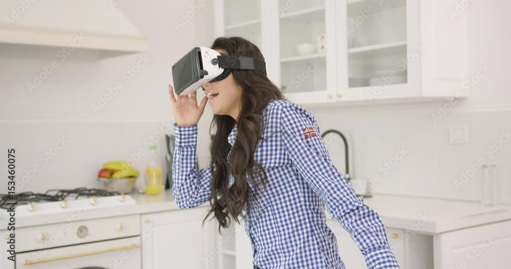 Beautiful young brunette in checkered shirt looking amazed while wearing goggles of virtual reality and standing in modern light kitchen. Beautiful young brunette in checkered shirt looking amazed while wearing goggles of virtual reality and standing in modern light kitchen.