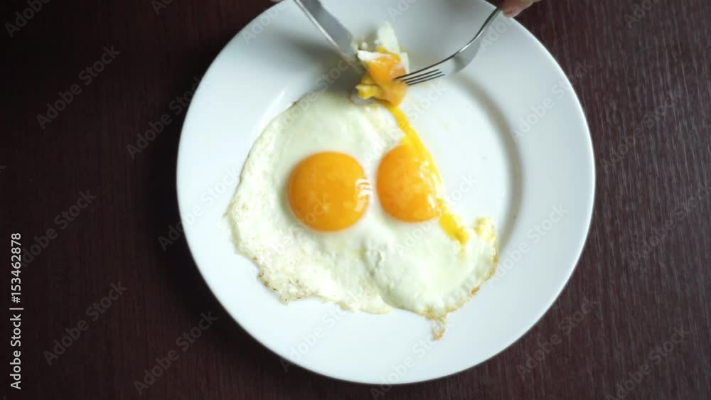 Cutting fried eggs with knife and fork on white plate at wooden table. Eating eggs. Traditional breakfast. Eating fried eggs with knife and fork. Top view of fried eggs on white plate Cutting fried eggs with knife and fork on white plate at wooden table. Eating eggs. Traditional breakfast. Eating fried eggs with knife and fork. Top view of fried eggs on white plate