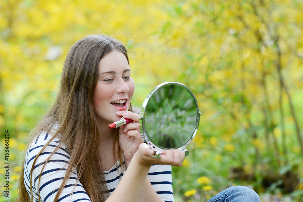 Beautiful young woman applying red lipstick looking at mirror isolated on natural background. Makeup and cosmetics concept. Beautiful young woman applying red lipstick looking at mirror isolated on natural background. Makeup and cosmetics concept.