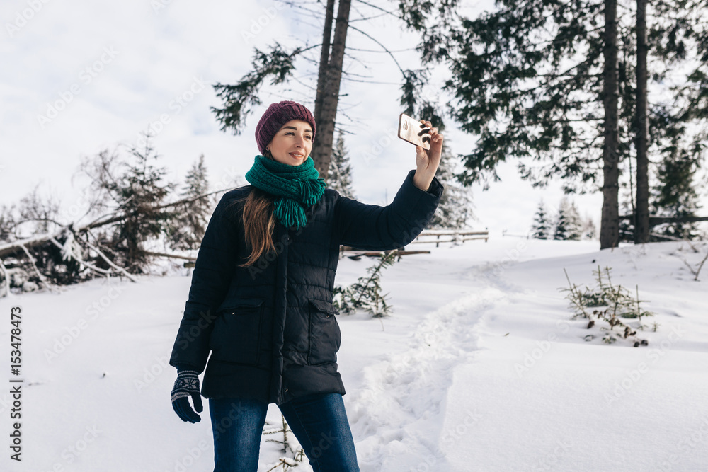 The pretty girl takes a selfie against the background of a landscape. On the street winter. On the girl a warm blue jacket, a lilac cap and a menthol scarf.