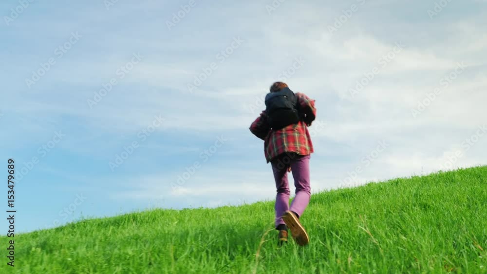 A young man with a backpack behind him rises to the top of a large green hill. In a beautiful place against the blue sky A young man with a backpack behind him rises to the top of a large green hill. In a beautiful place against the blue sky