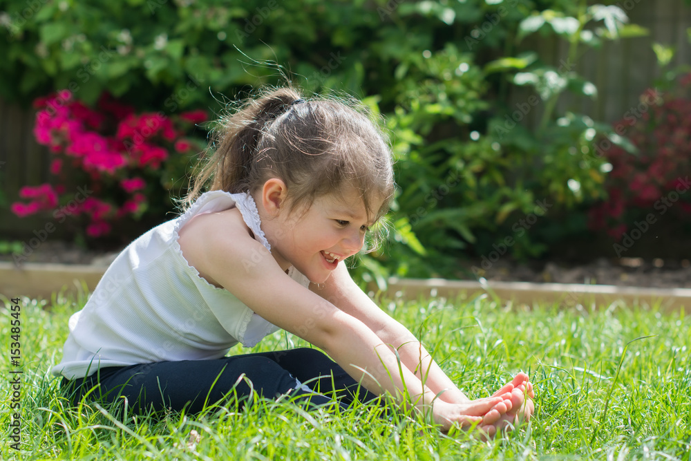 © Liliya Trott - Little girl sitting on the grass in the park doing yoga, reaching her toes, selective focus