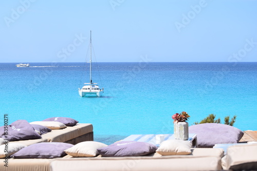 Luxuriöse blaue Yacht ankert im türkisfarbenen Wasser vor der Küste von Formentera, im Vordergrund Bänke und Kissen am Ufer, im Hintergrund ein Motorboot photo