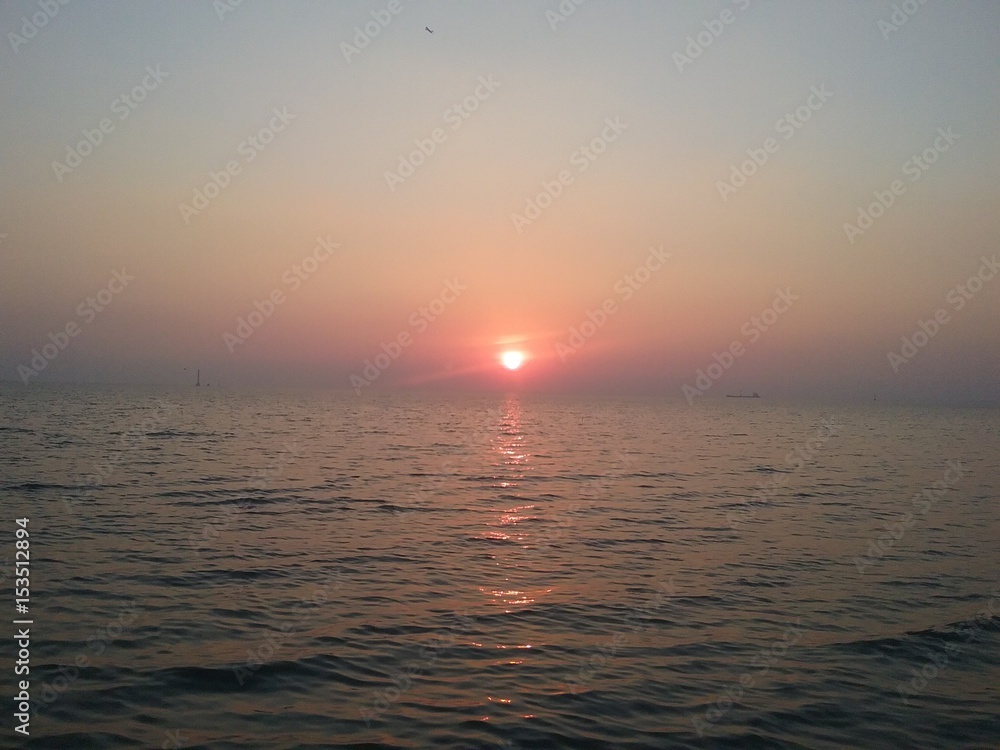 A blurred disk of bright sun over horizon against the background of a reddish sky. Sea sunset with reflection at water.