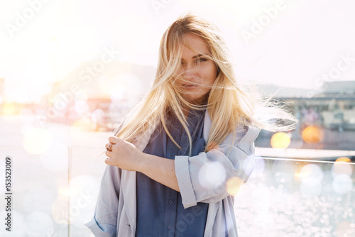 A portrait of a beautiful caucasian female touching her long blonde hair tumbled by wind while standing on a blurred city river background. A photo of a business woman having break. Flare lights.Bokeh