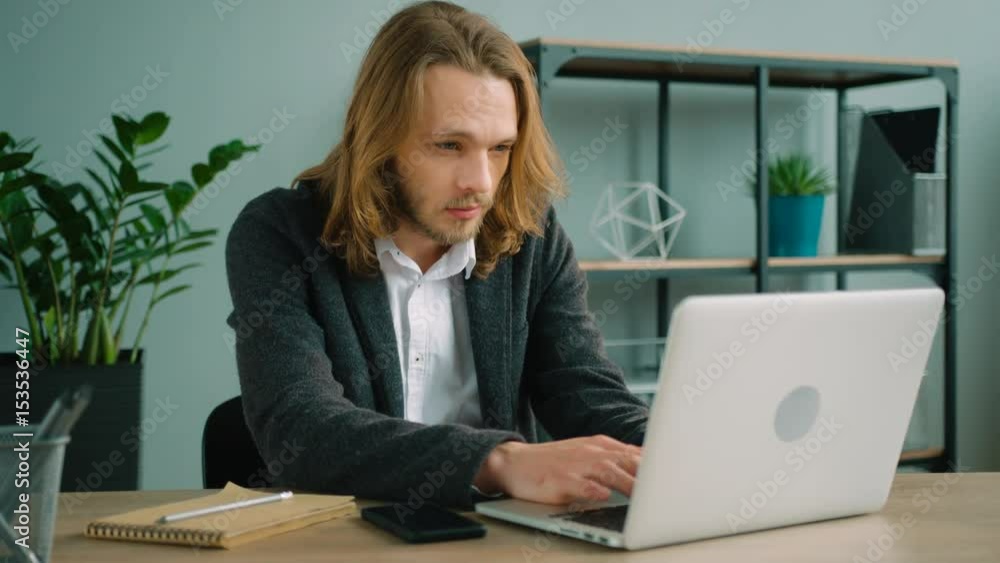 Young man sitting in the office and typing on his laptop computer. Long hair hipster using laptop pc, looking at screen while sitting at the office table. Young man sitting in the office and typing on his laptop computer. Long hair hipster using laptop pc, looking at screen while sitting at the office table.