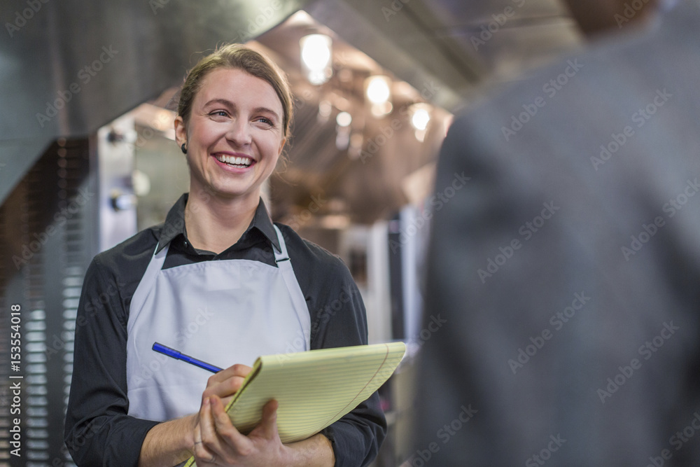 Smiling female chef with note pad talking with restaurant owner in kitchen Smiling female chef with note pad talking with restaurant owner in kitchen