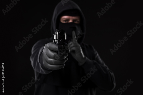 Fototapeta Masked robber with gun aiming into the camera against a black background