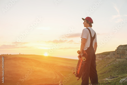 A guy in a cap and kabenison with a long board in his hands is standing in the nature against the sunset