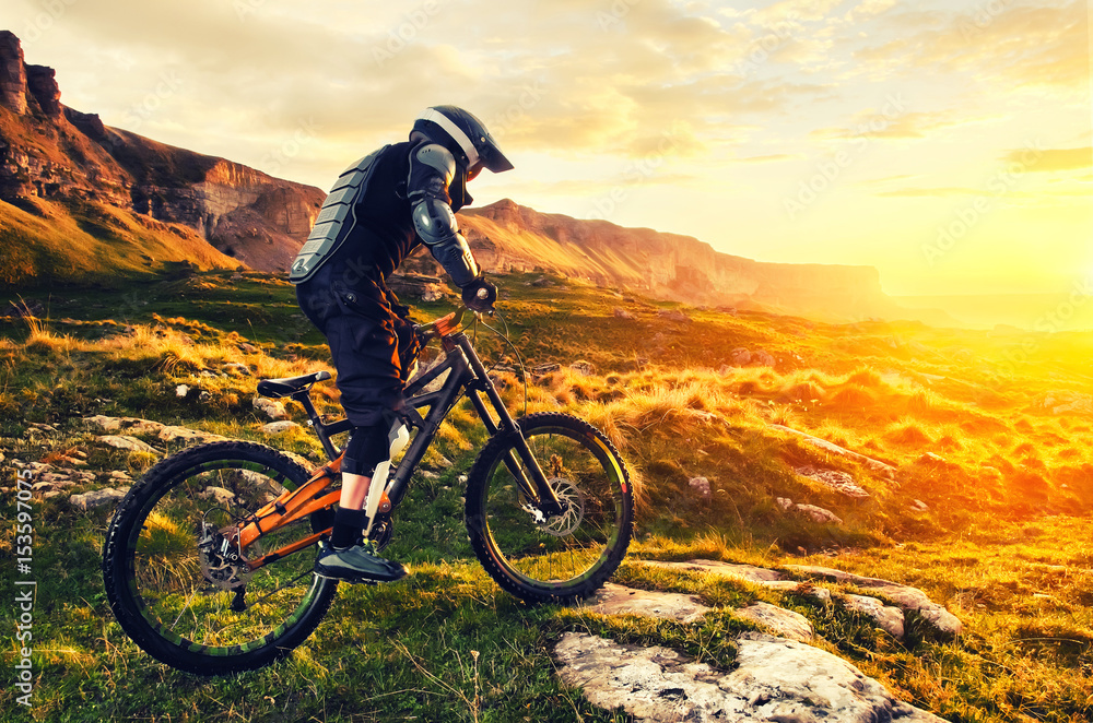 The rider in full protective equipment on the mtb bike is riding toward the sunset in the rays of the sunset sun against the background of the rocks of the setting sun and clouds