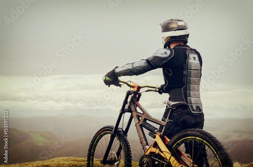 The rider in the full-face helmet and full protective equipment on the mtb bike stands on a rock against the background of a ridge and low clouds