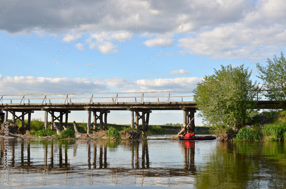 Man on kayak paddling under the pillars of old wooden bridge.