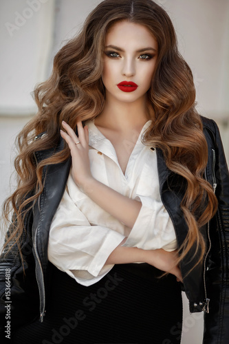 Fashion outdoor portrait of gorgeous long hair woman in elegant red dress and black leather jacket - autumn style. Fashionable hipster girl in trendy clothes posing at city street lifestyle portrait.