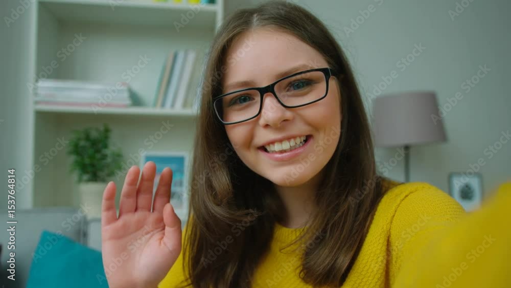 Beautiful young teen girl video chatting, waving hand, blogging in the living room at home. Girl smiling to the camera. Close up Beautiful young teen girl video chatting, waving hand, blogging in the living room at home. Girl smiling to the camera. Close up
