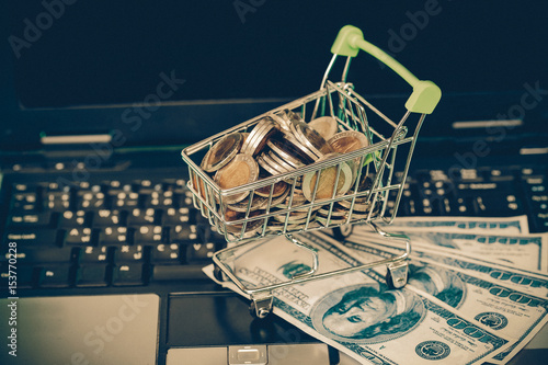 Small shopping cart contains coins on laptop and working space on wooden .