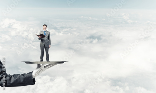 Businessman on metal tray with red book in hands against blue sky background