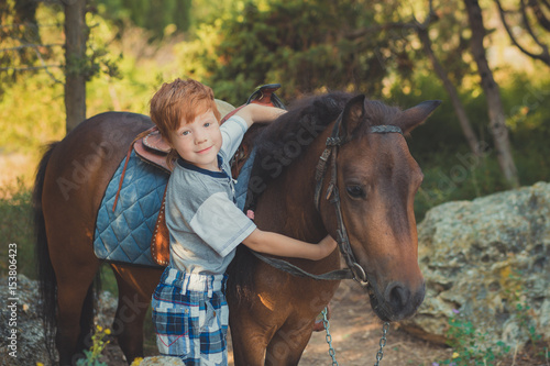 Cute red hair boy smiling to camera and standing in forest with horse