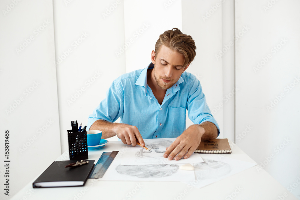Young handsome confident pensive businessman sitting at table with pencil drawing portrait. White modern office interior background.