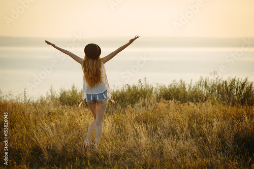 Young pretty woman wearing hat and sunglasses dressed in boho style with open arms under the sunrise on the nature near the sea