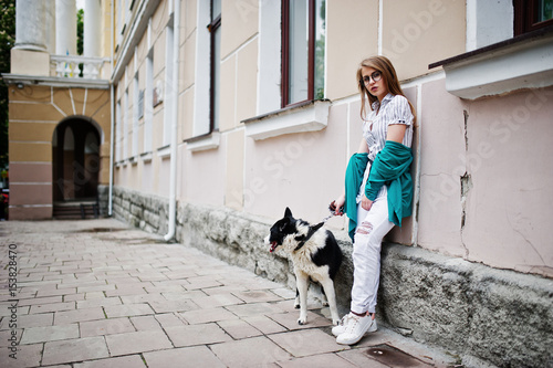 Trendy girl at glasses and ripped jeans with russo-european laika (husky) dog on a leash, against street of city. Friend human with animal theme.