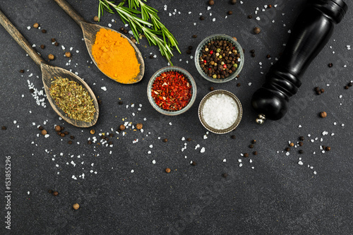On a black stone surface of spices, black pepper mill, in wooden spoons of turmeric, dried basil, in glass bowls pepper peas, salt, paprika, rosemary, colorful