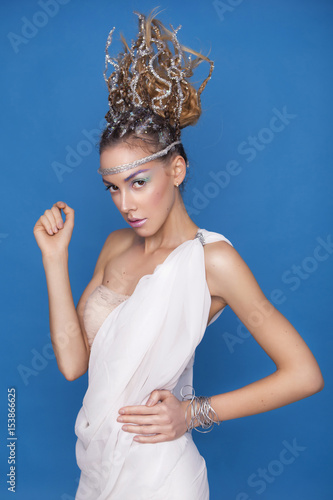 Beautiful brunette blonde young skiny woman in greece mythology styled outfit white toga and creative abstract hairstyle and creative make up on a neutral blue background. Glowing clean fresh skin