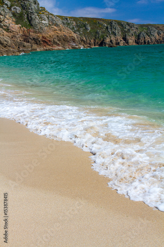 The sandy beach and emerald green ocean off Porthcurno beach in Cornwall, UK with cliffs in the background and white waves or surf crashing over the sand.
