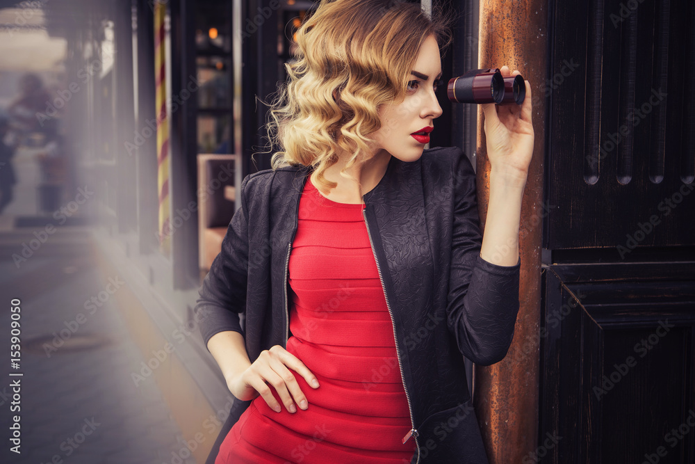 Beautiful brunette sexy spy agent (killer or police) woman in leather jacket and red luxury dress with a binocular in her hand stalking someone, to catch him in european city Stock Photo  image
