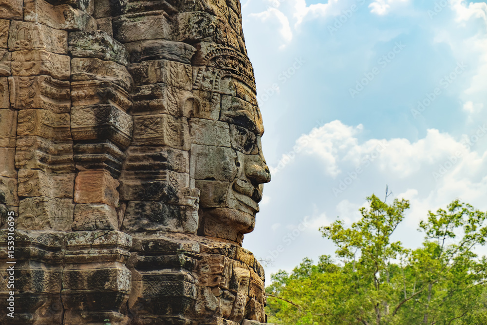 Obraz premium Smiling stone face of Prasat Bayon, the central temple of Angkor Thom Complex, Siem Reap, Cambodia. Ancient Khmer temple is surrounded by tropical trees, famous Cambodian landmark, World Heritage.