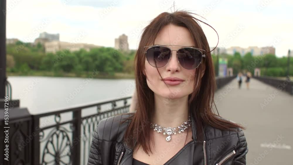 Beautiful brunette in a leather jacket walks the bridge and puts on sunglasses. Girl moves slowly and looks at the camera. Beautiful brunette in a leather jacket walks the bridge and puts on sunglasses. Girl moves slowly and looks at the camera.