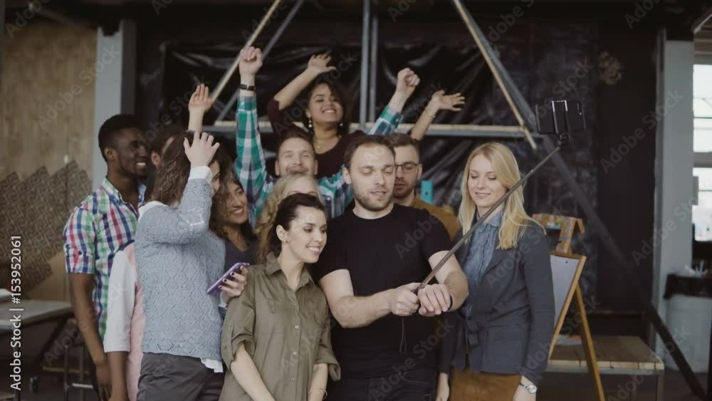 Happy team takes selfie at loft office. Man holds smartphone with self-stick and takes photo of workers, smiling friends Happy team takes selfie at loft office. Man holds smartphone with self-stick and takes photo of workers, smiling friends