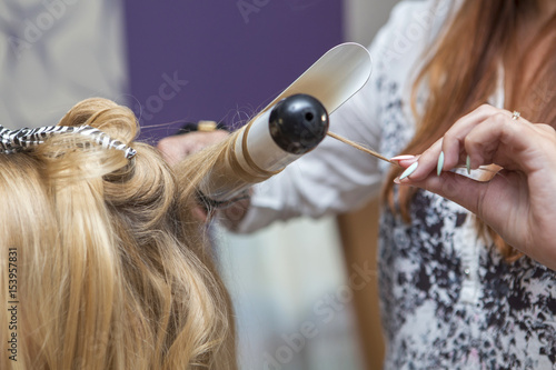 Brunette red hair hairdresser artist making curly hairstyle to blonde bride woman on her wedding day with iron. Studio interior, close up of a hands in process