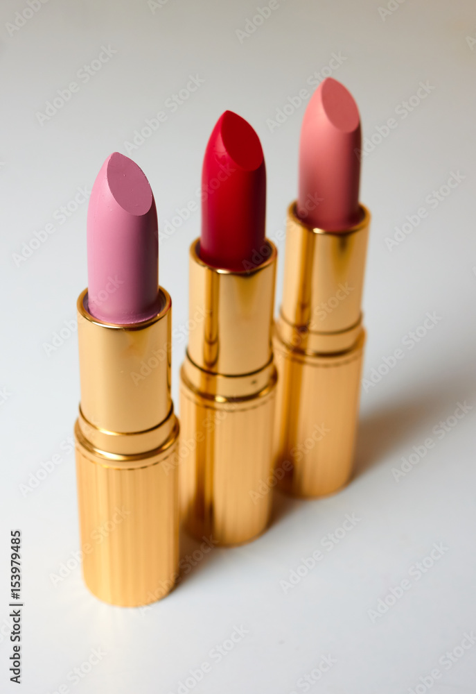 Three matte lipsticks in luxurious gold tubes stand on a white surface. Vertical photo. Red lipstick, pink and lipstick flesh-colored Three matte lipsticks in luxurious gold tubes stand on a white surface. Vertical photo. Red lipstick, pink and lipstick flesh-colored