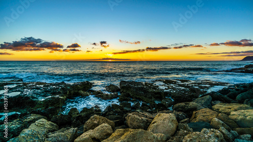 Sunset over the horizon with a few clouds and waves crashing on the rocky shores of the west coast of the tropical Hawaiian island of Oahu photo