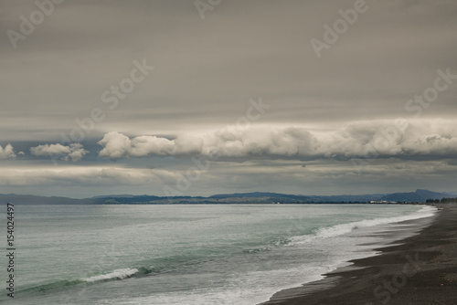 Fototapeta Naklejka Na Ścianę i Meble -  Napier, New Zealand - March 9, 2017: South side of Hawkes Bay with black lava beach and surf in front, all under heavy, dark storm clouds cut by band of white clouds. Shine on Pacific Ocean water.