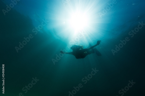A young woman elegantly free dives into the depths of the Pacific Ocean as the sun illuminates the water around her.