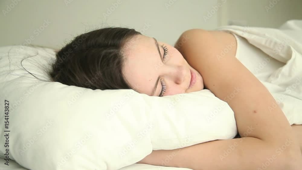 Young beautiful happy female sleeping soundly on bed in bedroom at home or hotel, smiling in her dream, hugging white comfy soft pillow, lying with her eyes closed on cozy mattress in relaxed pose Young beautiful happy female sleeping soundly on bed in bedroom at home or hotel, smiling in her dream, hugging white comfy soft pillow, lying with her eyes closed on cozy mattress in relaxed pose