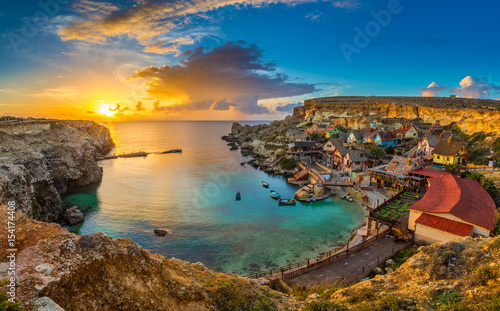 Il-Mellieha, Malta - Panoramic skyline view of the famous Popeye Village at Anchor Bay at sunset with traditional Luzzu boats, beautiful colorful clouds and sky photo