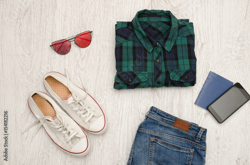 Blue-green checkered shirt, glasses, sneakers, jeans, phone and passport. Wooden background. Fashionable concept, top view