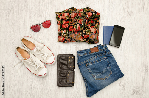 Shirt in flowers, glasses, sneakers, jeans, phone and passport. Wooden background. Fashionable concept, top view