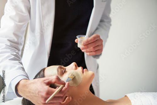 Beautician putting mask on young female face with brush