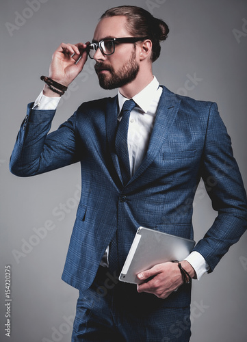 portrait of handsome fashion businessman  model dressed in elegant blue suit on gray background in studio. Touching his glasses and using touchpad photo