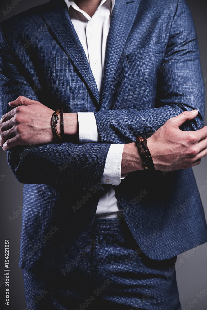 Fototapeta premium Fashion portrait of young businessman handsome model man dressed in elegant blue suit with accessories on hands posing on gray background in studio. Keeping crossed hands