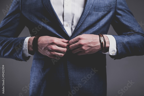 Fashion portrait of young businessman handsome model man dressed in elegant blue suit with accessories on hands posing on gray background in studio. Buttoning his jacket