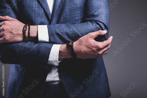 Fashion portrait of young businessman handsome model man dressed in elegant blue suit with accessories on hands posing on gray background in studio. Keeping crossed hands
