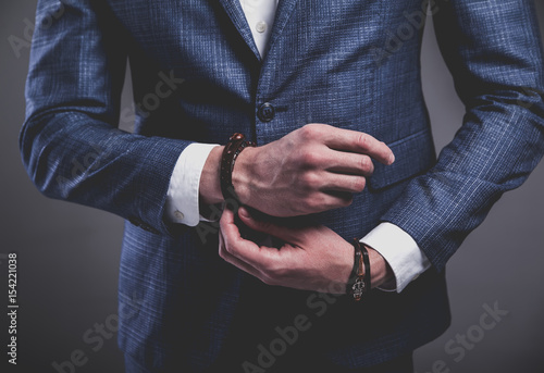 Fashion portrait of young businessman handsome model man dressed in elegant blue suit with accessories on hands posing on gray background in studio. Buttoning his jacket