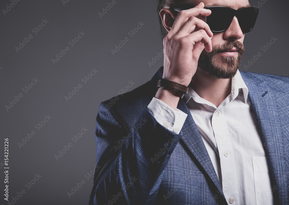 portrait of handsome fashion stylish hipster businessman model dressed in elegant blue suit posing on gray background in studio. Touching his sunglasses
