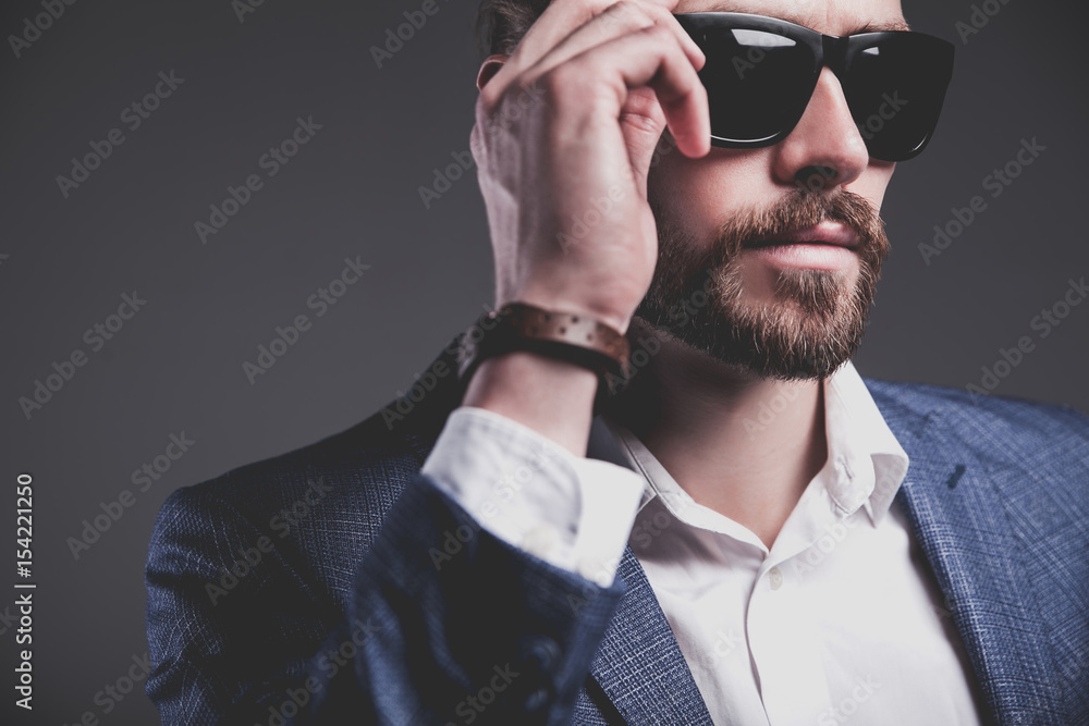 Obraz premium portret przystojny moda model stylowy hipster biznesmen ubrany w elegancki niebieski garnitur pozowanie na szarym tle w studio. Dotykając jego okularów przeciwsłonecznych