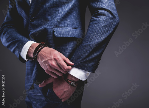 Fashion portrait of young businessman handsome model man dressed in elegant blue suit with accessories on hands posing on gray background in studio. Hands in pockets